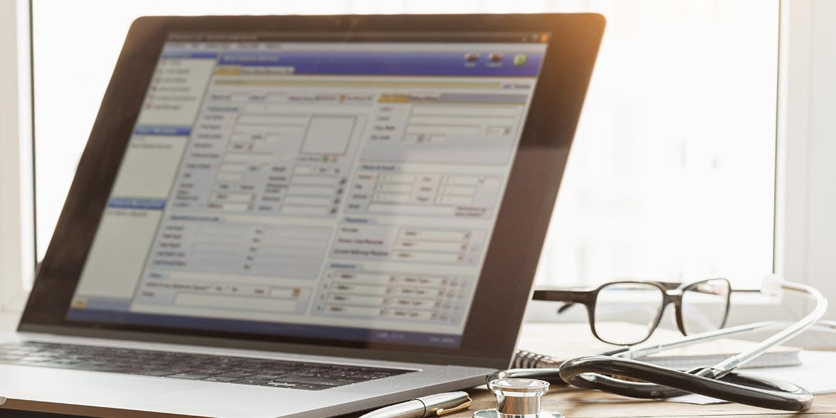 A laptop displaying an electronic health record (EHR) system sits on a wooden desk next to a stethoscope, glasses, and a pen. The screen is visible, potentially exposing sensitive patient information, highlighting a possible HIPAA violation.